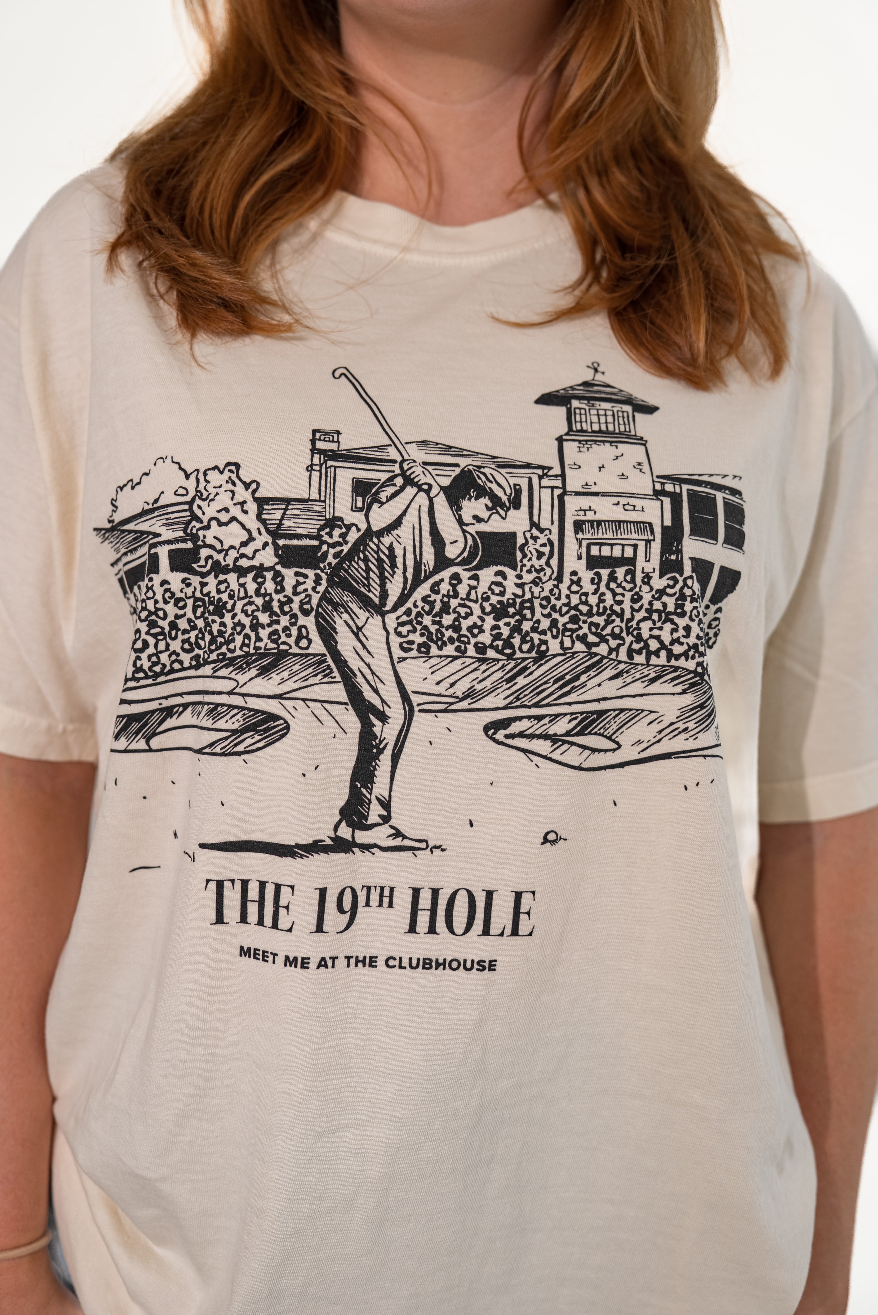Close-up of the front of a cream-colored t-shirt featuring a detailed black graphic of a golfer taking a swing outside a clubhouse, with the text 