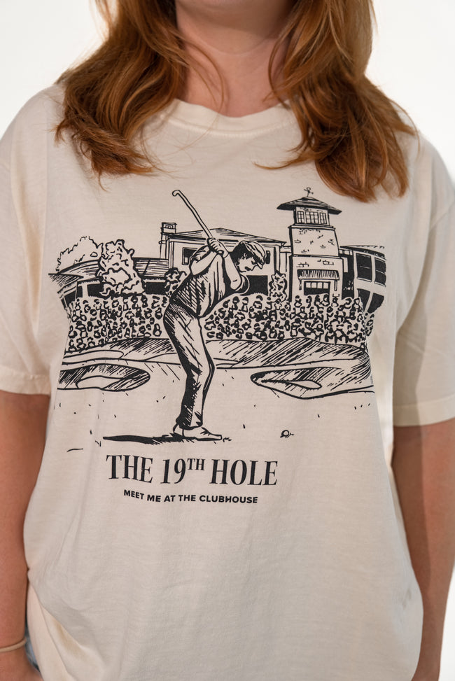 Close-up of the front of a cream-colored t-shirt featuring a detailed black graphic of a golfer taking a swing outside a clubhouse, with the text 