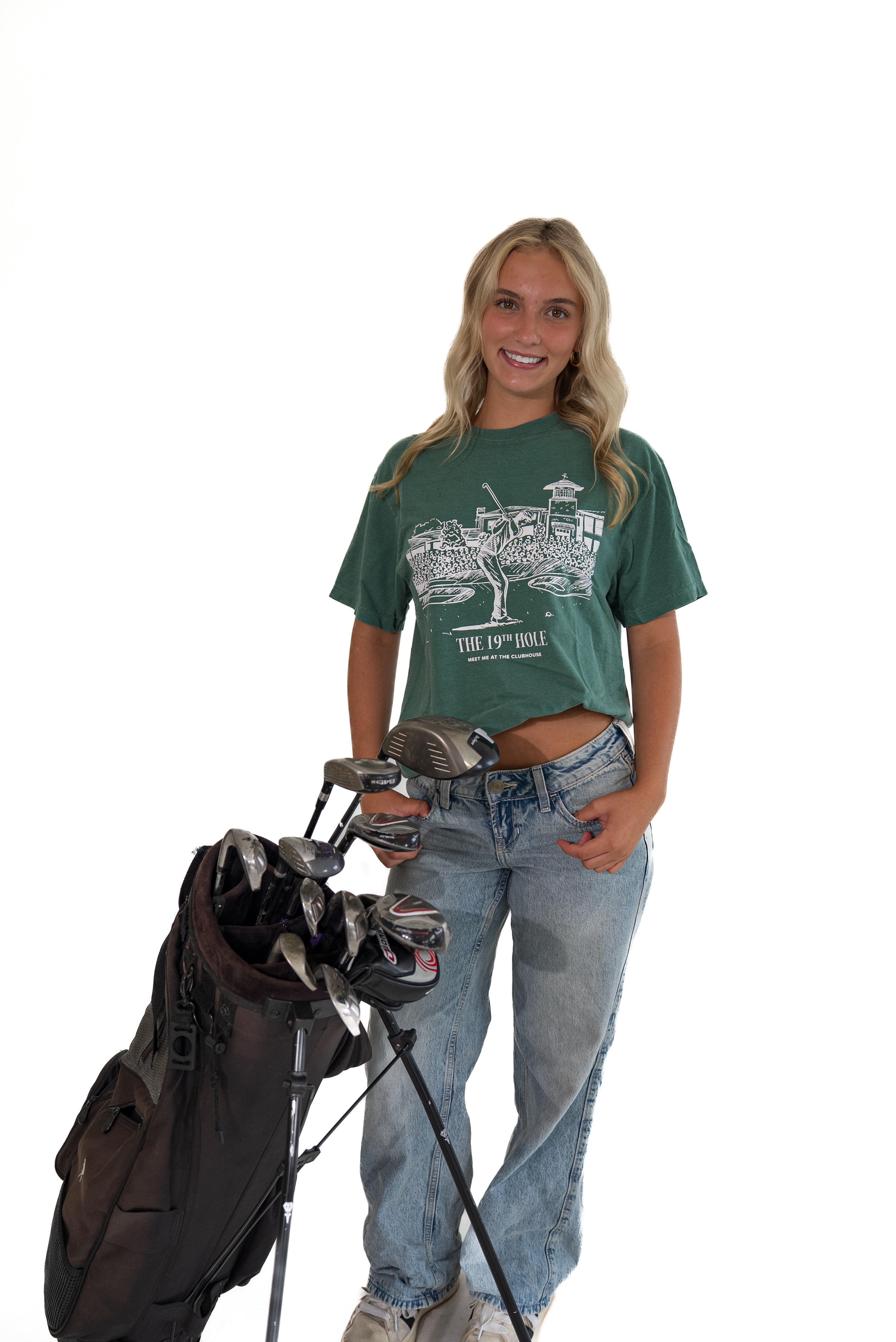 Smiling woman standing against a white background next to a golf bag, wearing a green short-sleeve graphic t-shirt with a white golf illustration, and light-wash blue jeans.