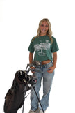 Smiling woman standing against a white background next to a golf bag, wearing a green short-sleeve graphic t-shirt with a white golf illustration, and light-wash blue jeans.