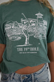 Close-up of the front of a green t-shirt featuring a white graphic of a golfer taking a swing in front of a clubhouse, with the text 