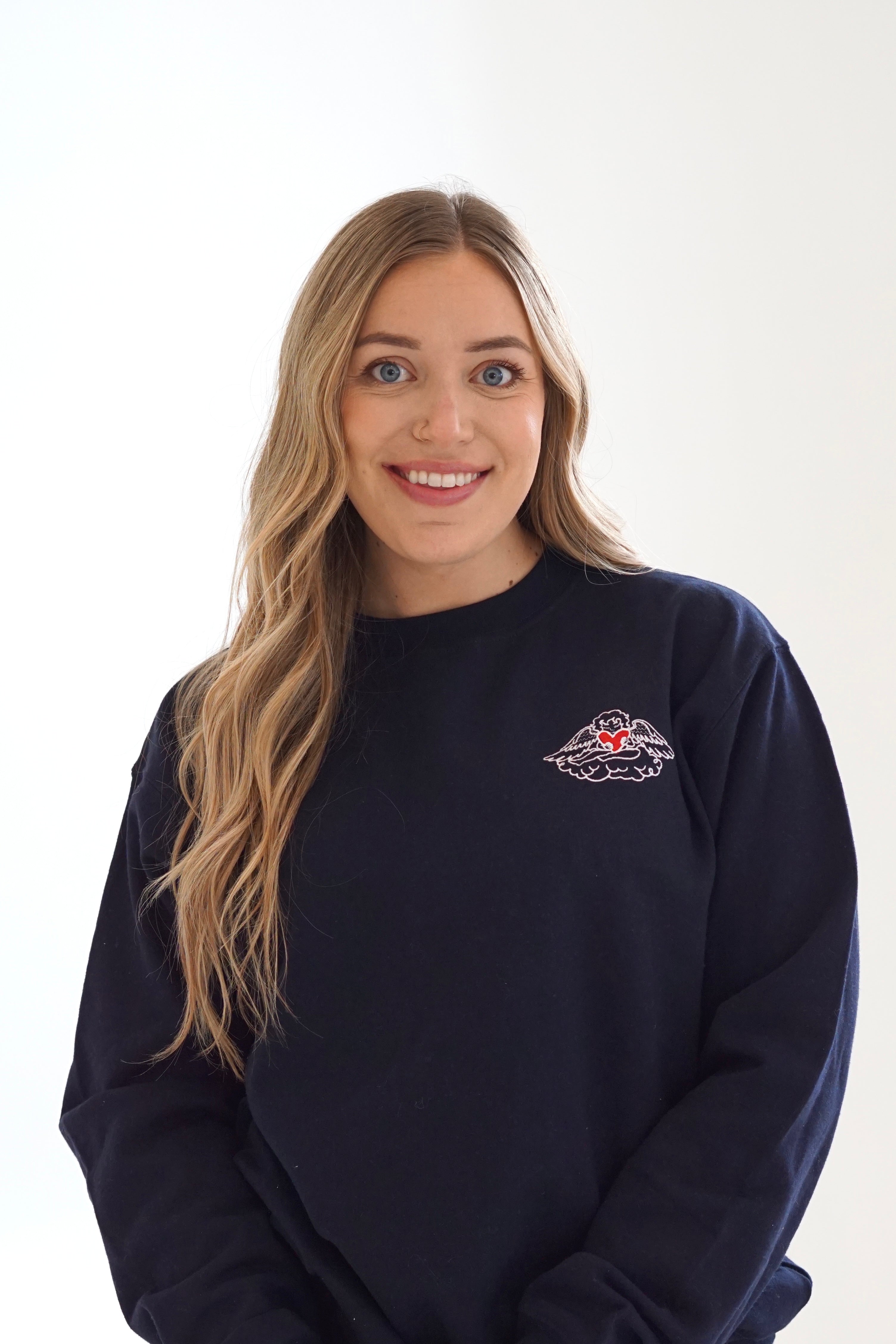 Woman wearing a black Angel Heart crewneck sweatshirt with a red heart and wing embroidered graphic on the chest.