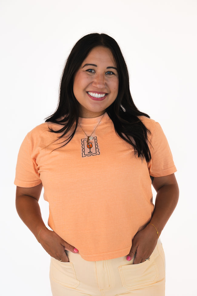Woman wearing a peach fitted baby tee with a centered embroidered postage stamp design featuring a cocktail glass.