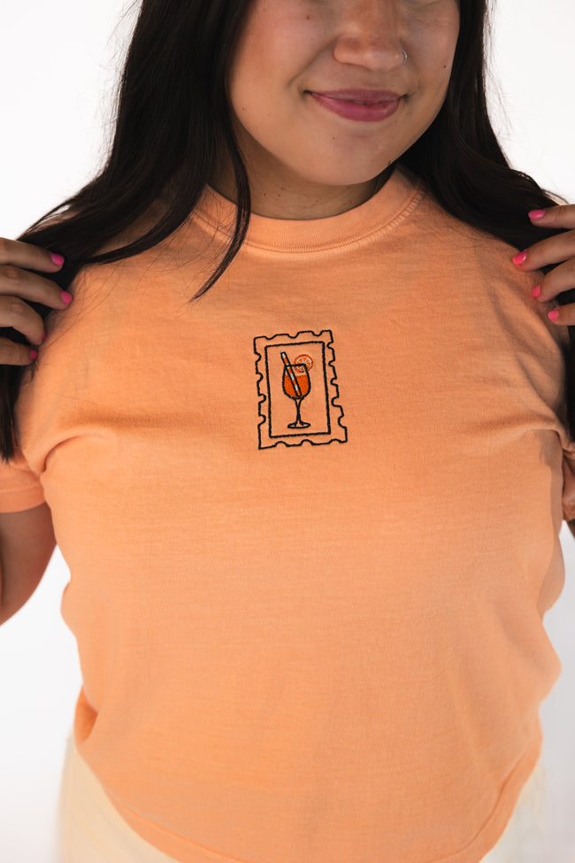 Woman wearing a peach fitted baby tee with a centered embroidered postage stamp design featuring a cocktail glass.