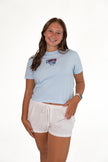 Smiling woman standing against a white background, wearing a light-blue short-sleeve graphic tee with a patriotic USA popsicle patch, white drawstring lounge shorts, and red sandals.