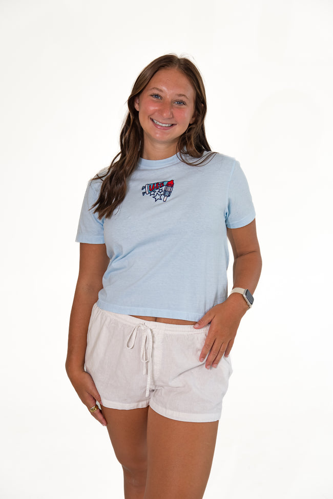 Smiling woman standing against a white background, wearing a light-blue short-sleeve graphic tee with a patriotic USA popsicle patch, white drawstring lounge shorts, and red sandals.