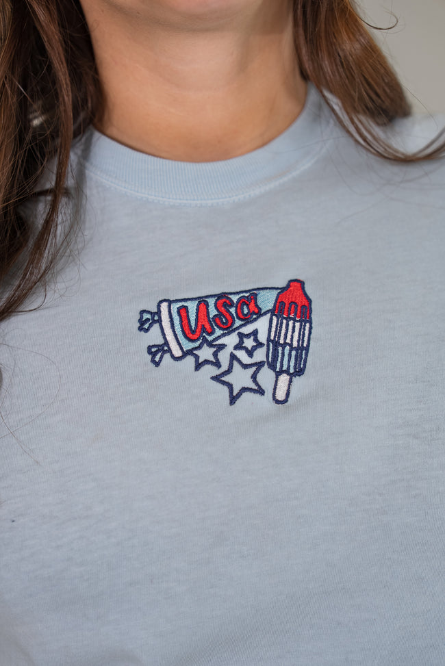 Close-up of the front of a light-blue t-shirt featuring a detailed embroidered graphic of a patriotic rocket popsicle and a 