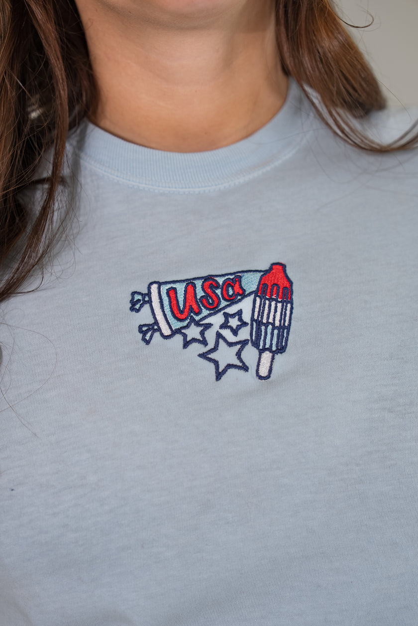 Close-up of the front of a light-blue t-shirt featuring a detailed embroidered graphic of a patriotic rocket popsicle and a 
