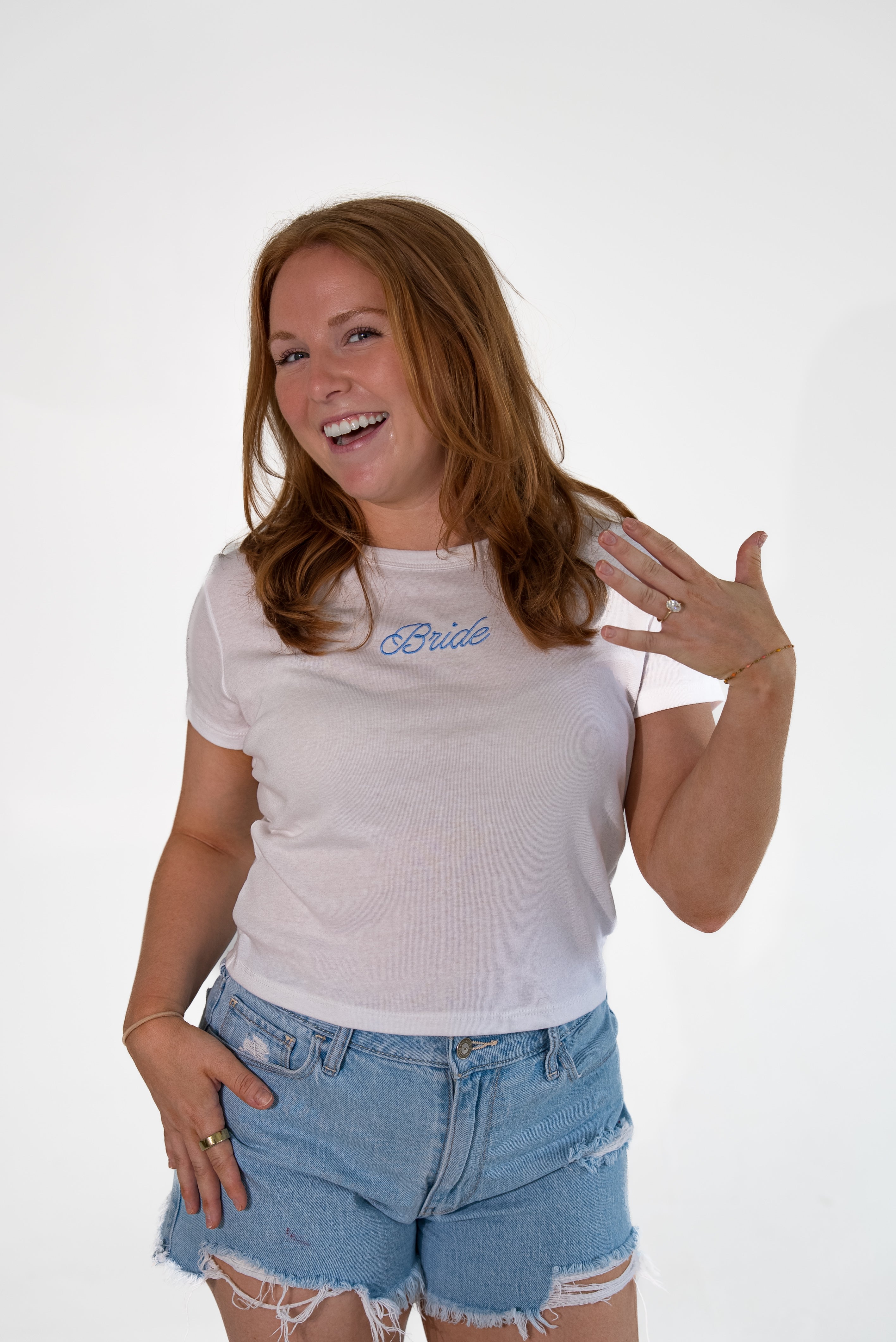A woman with long light brown hair smiling and posing in a white t-shirt with 'Bride' printed in blue cursive on the chest, paired with light-wash distressed denim shorts, against a plain white studio background.