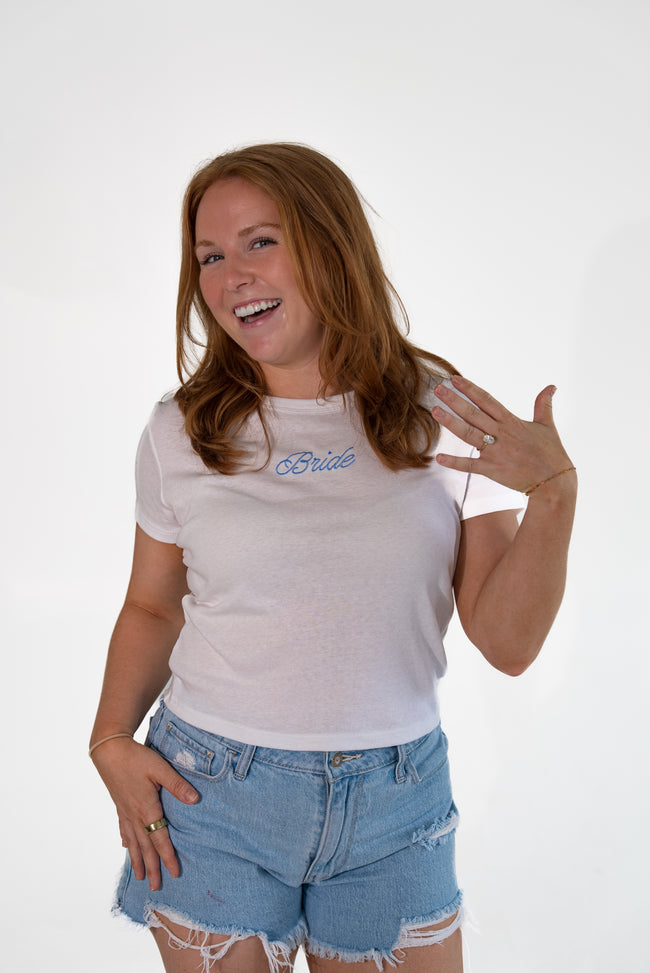 A woman with long light brown hair smiling and posing in a white t-shirt with 'Bride' printed in blue cursive on the chest, paired with light-wash distressed denim shorts, against a plain white studio background.