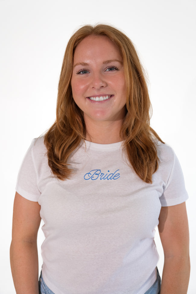A smiling woman with long light brown hair wearing a white crewneck t-shirt with the word 'Bride' embroidered in blue script across the chest, set against a plain white background.