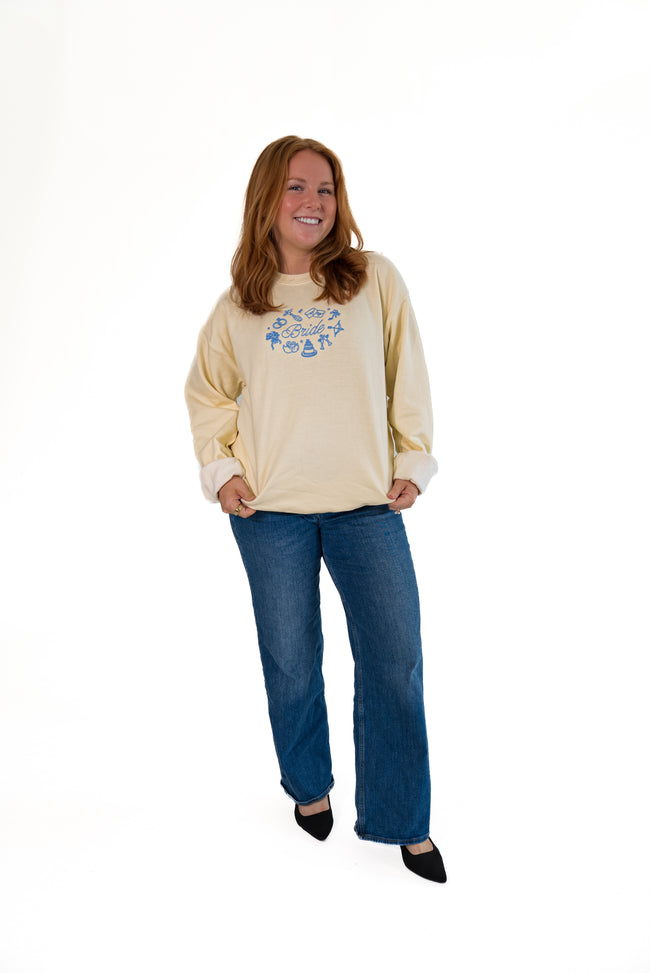 A woman modeling a cream-colored sweatshirt with a blue graphic on the chest, paired with blue jeans and black shoes, against a plain white background.