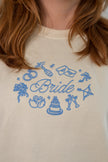 Close-up of a person wearing a cream-colored sweatshirt with a blue embroidered graphic that reads 'Bride' surrounded by wedding-themed icons like rings, a cake, and champagne glasses.