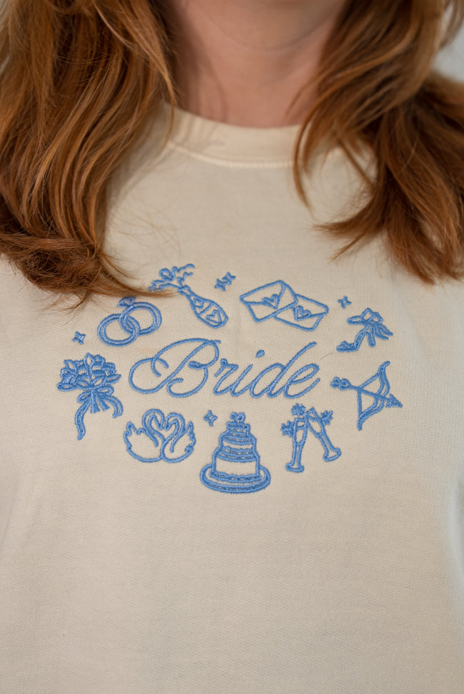 Close-up of a person wearing a cream-colored sweatshirt with a blue embroidered graphic that reads 'Bride' surrounded by wedding-themed icons like rings, a cake, and champagne glasses.