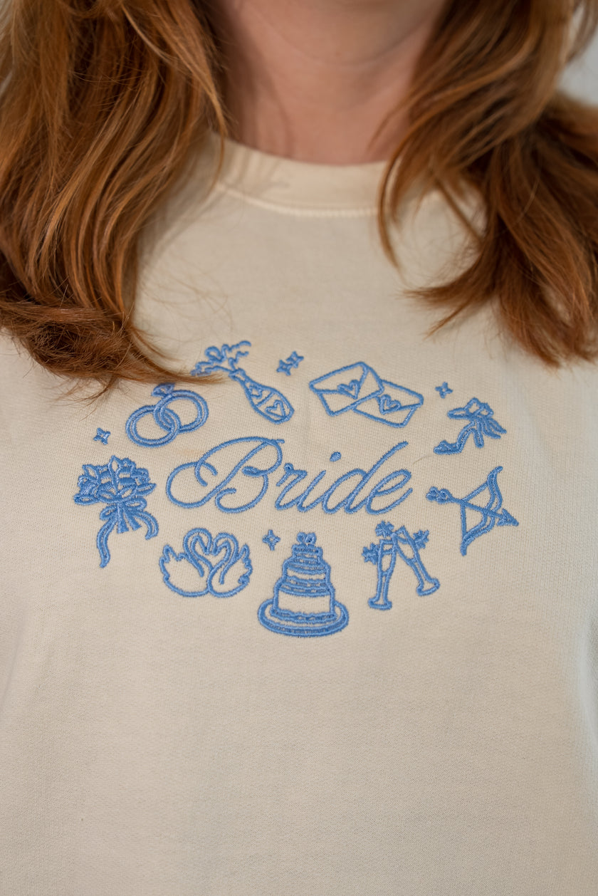 Close-up of a person wearing a cream-colored sweatshirt with a blue embroidered graphic that reads 'Bride' surrounded by wedding-themed icons like rings, a cake, and champagne glasses.