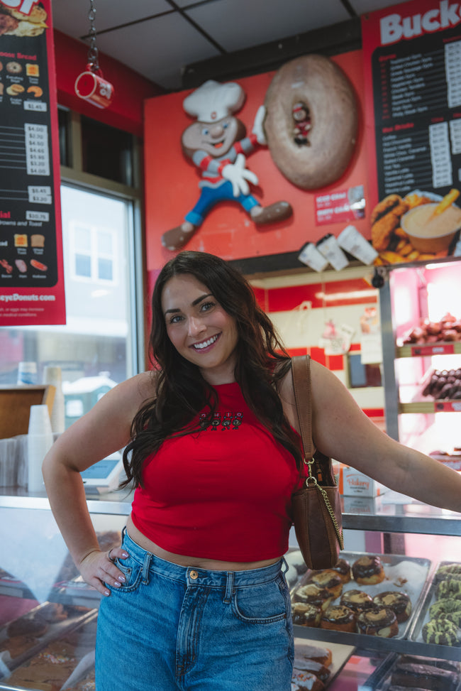 A woman posing inside a bakery or donut shop, wearing a bright red crop top and blue jeans, with a brown shoulder bag. The background features a red wall with a cartoon character and a large donut, along with menu boards and a glass display case of pastries.