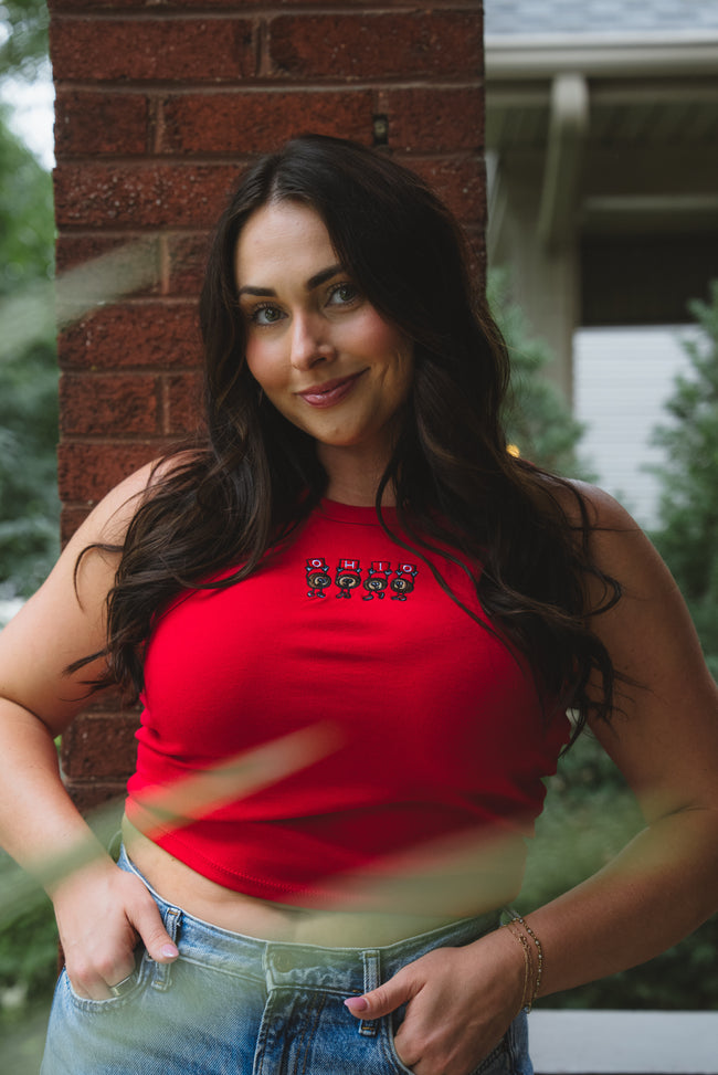 A woman with long dark hair wearing a bright red halter crop top with a small embroidered graphic on the chest, paired with blue jeans, standing outdoors in front of a brick pillar.