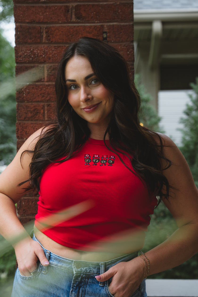 A woman with long dark hair wearing a bright red halter crop top with a small embroidered graphic on the chest, paired with blue jeans, standing outdoors in front of a brick pillar.