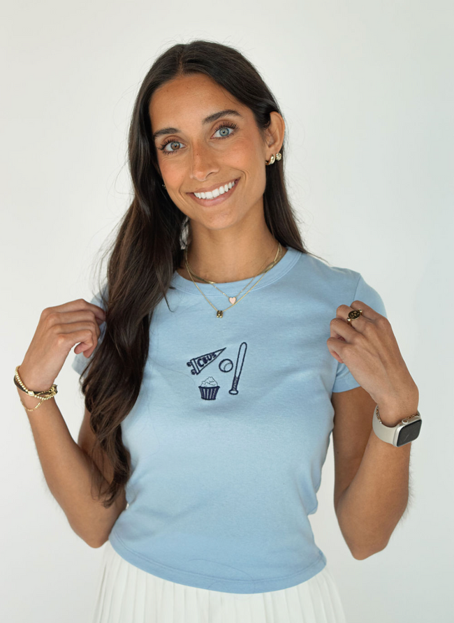 Woman wearing a light blue baby tee embroidered with a CBUS pennant, baseball bat, baseball, and popcorn design.