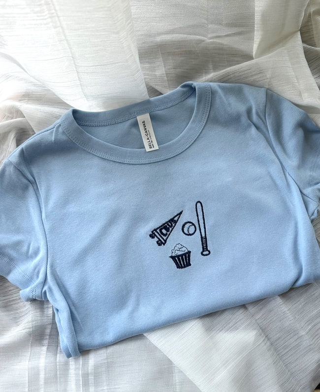 Flat lay of a light blue Columbus baseball baby tee with embroidered game day icons on the chest.