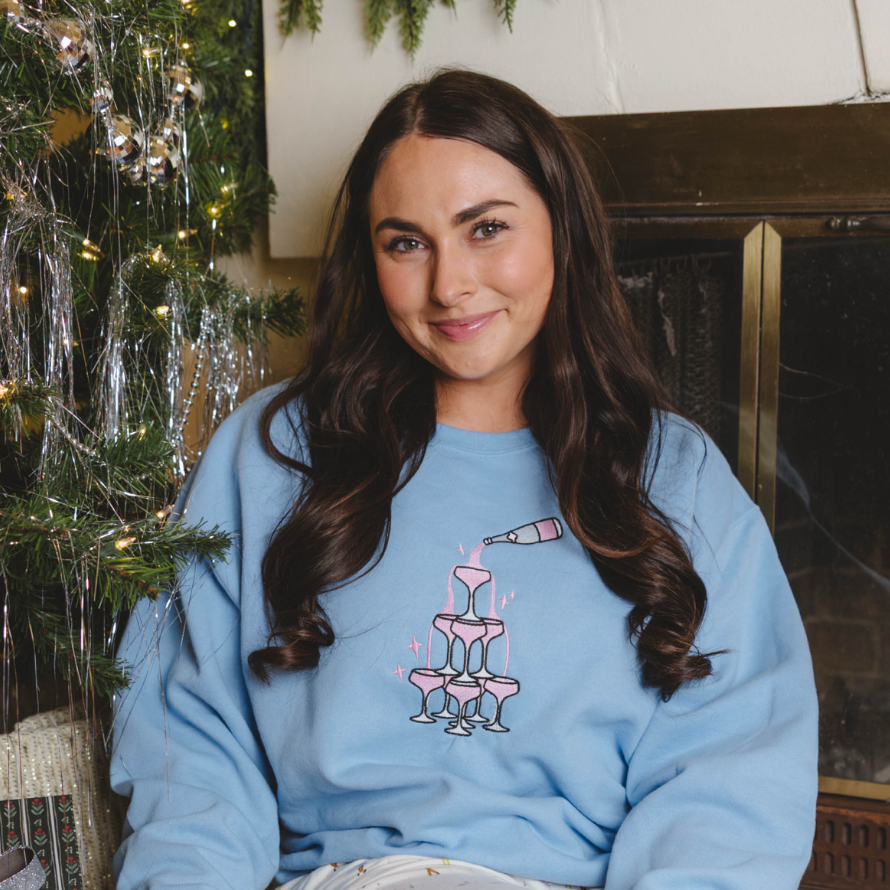Model wearing a light blue Champagne Tower graphic crewneck sweatshirt in a festive holiday setting.