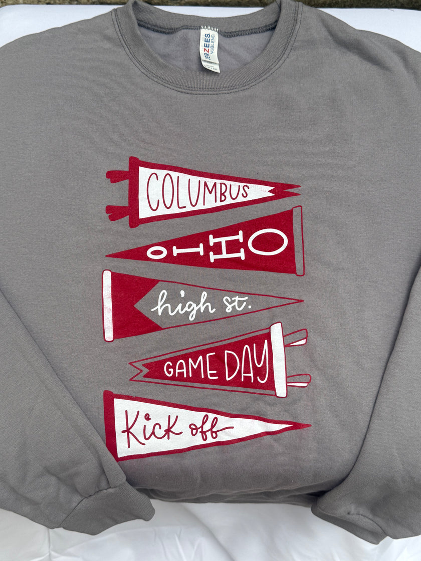 A gray crewneck sweatshirt featuring a graphic of five red and white pennants stacked vertically with the words 'COLUMBUS', 'OHIO', 'high st.', 'GAME DAY', and 'Kick off'.