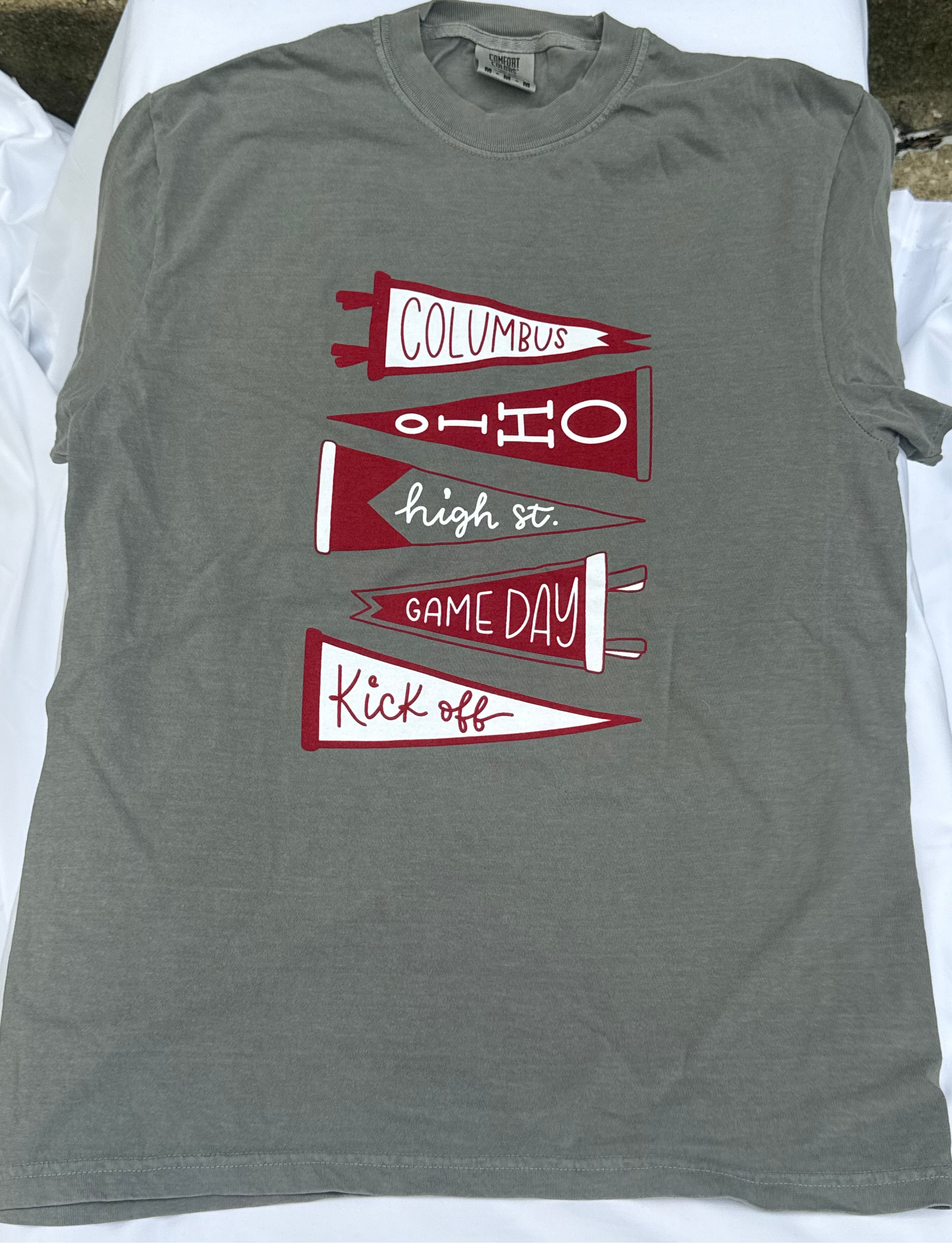 A heather gray short-sleeve t-shirt laid flat on white fabric, featuring a graphic of four red and white pennants with the words 'Columbus', 'Ohio', 'high st. GAME DAY', and 'Kick off'.