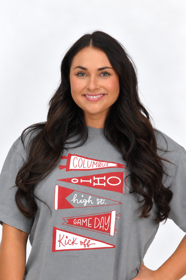 Woman wearing a gray graphic T-shirt featuring red and white pennant designs with “Columbus,” “Ohio,” “High St.,” “Game Day,” and “Kickoff” lettering.
