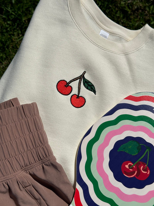 Flat lay of the Cherry Embroidered Crew Neck styled with brown shorts and a colorful cherry-themed plate.