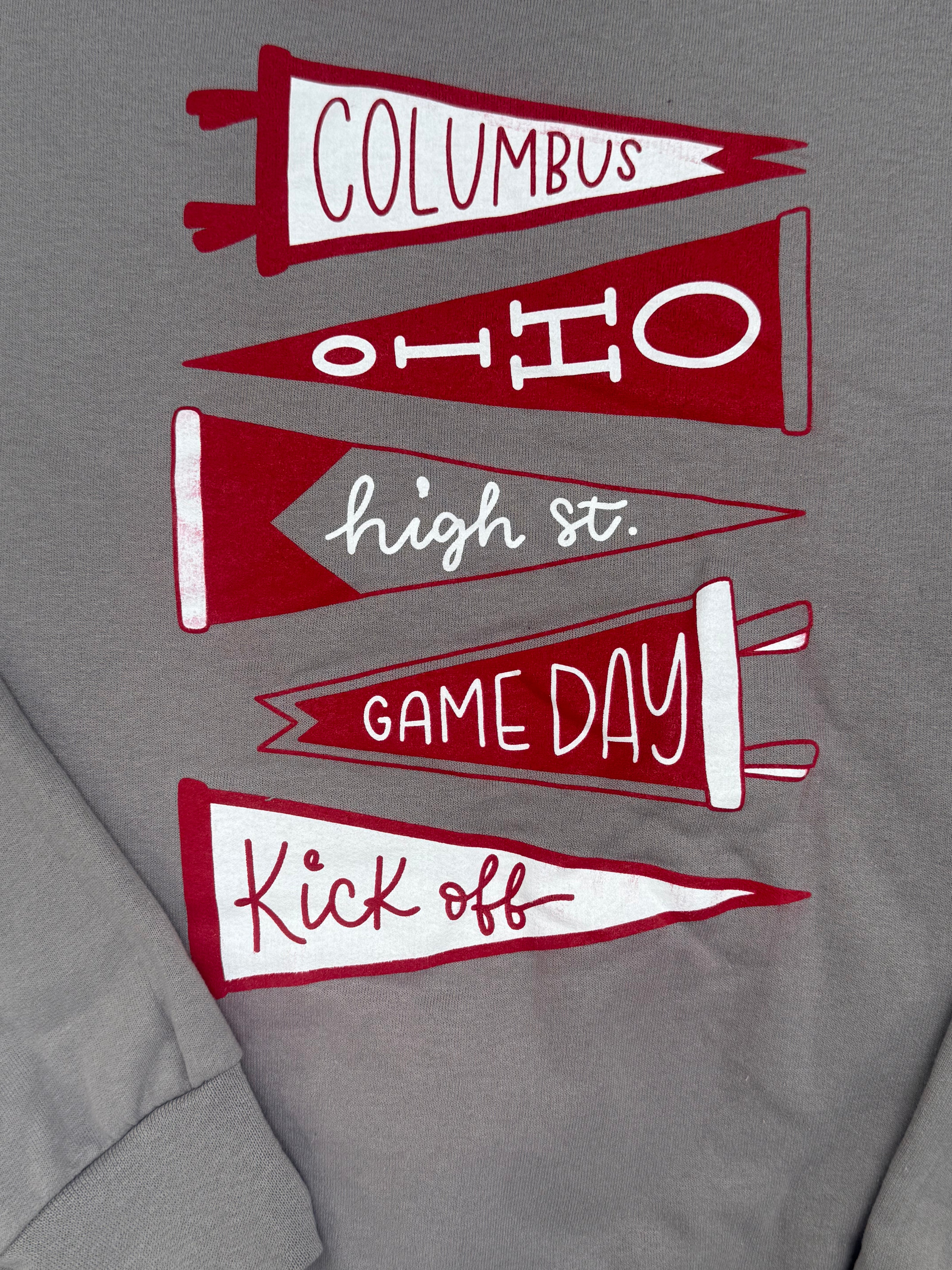 Close-up of a gray T-shirt featuring a graphic of five red and white pennants with text reading 'COLUMBUS', 'OHIO', 'high st.', 'GAME DAY', and 'Kick off'.