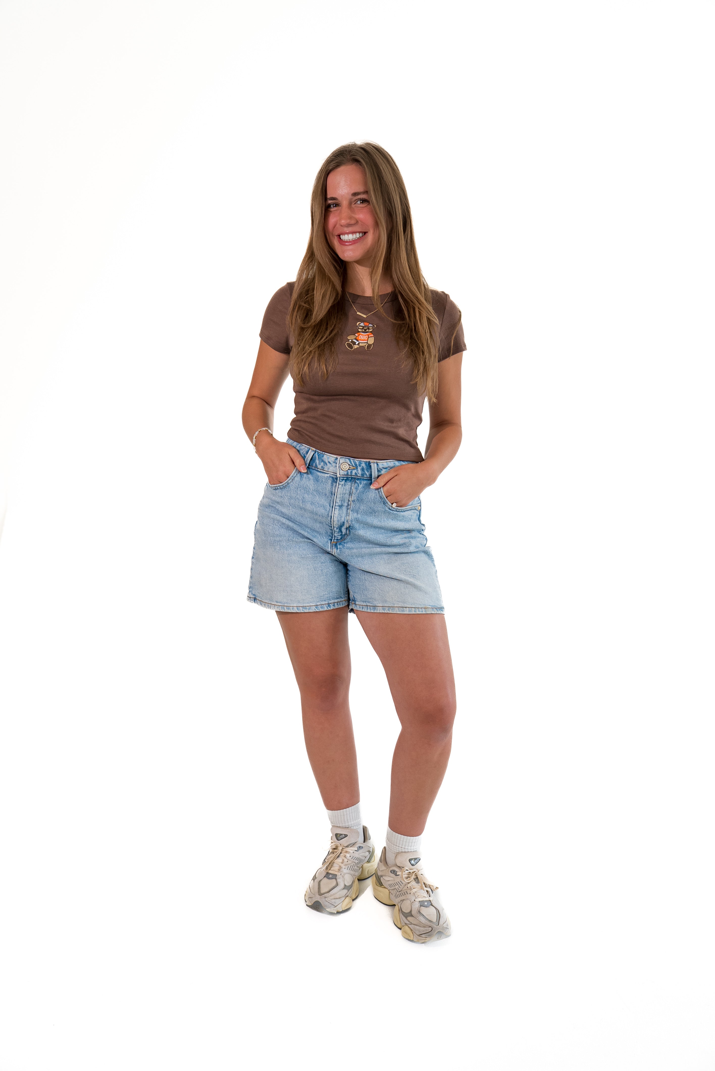 A woman modeling a brown short-sleeve t-shirt with a small graphic on the chest, paired with light-wash denim shorts, white socks, and beige sneakers, standing against a solid white background.