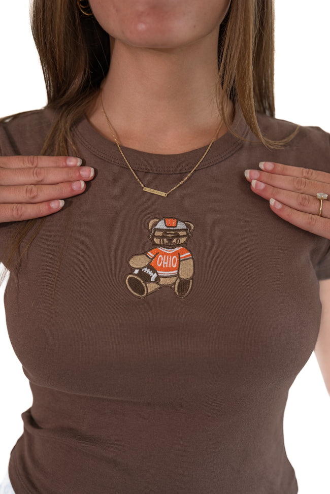 A close-up of a woman wearing a brown ribbed short-sleeve t-shirt with a teddy bear graphic on the chest. The graphic depicts a brown bear wearing an orange and white football helmet and jersey with 'OHIO' written on it.