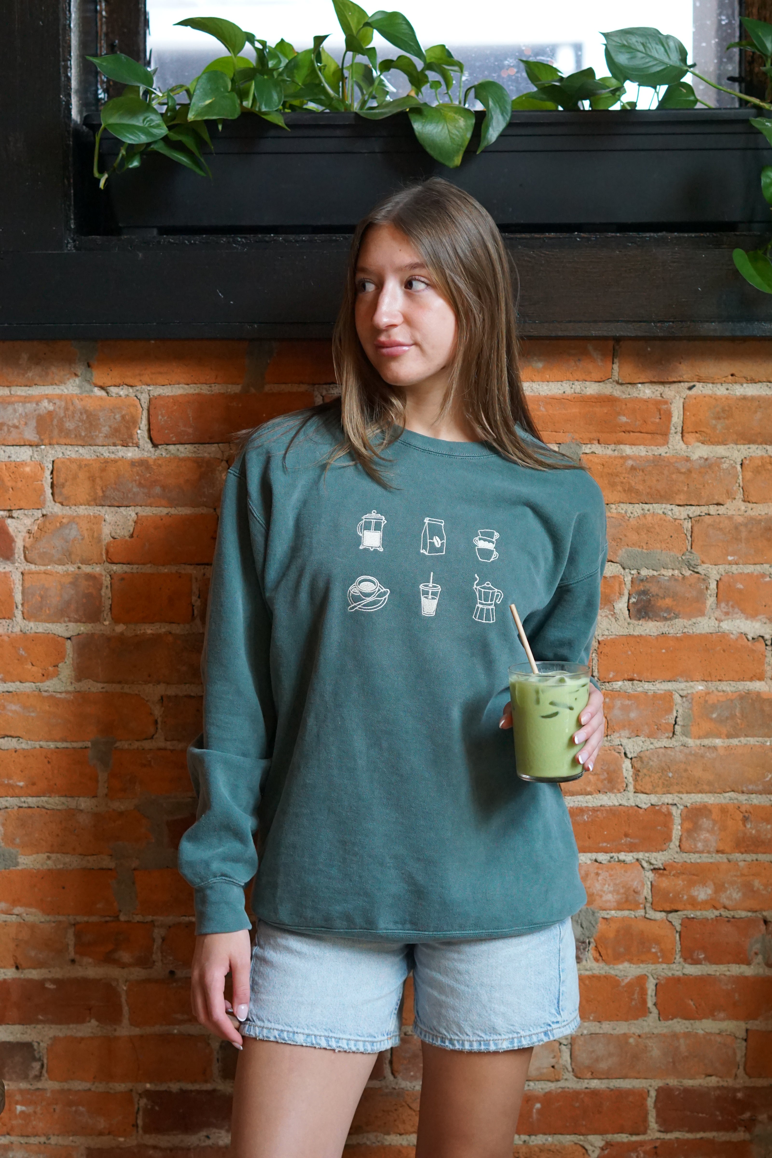 Woman wearing a forrest green crewneck sweatshirt with embroidered coffee icons.