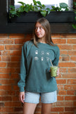 Woman wearing a forrest green crewneck sweatshirt with embroidered coffee icons.