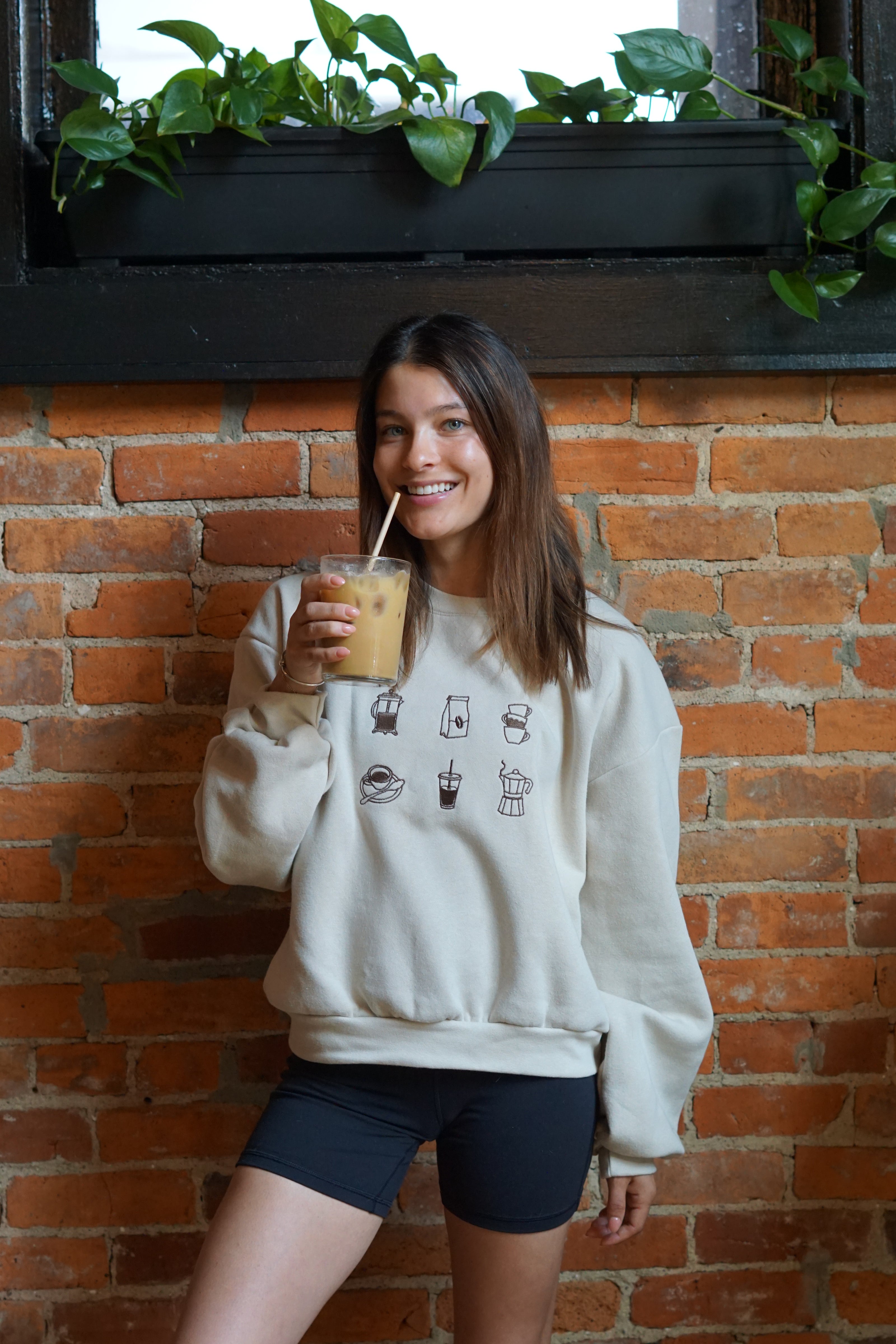 Woman wearing a cream crewneck sweatshirt embroidered with minimalist coffee icons while holding an iced coffee.