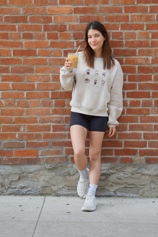 Cream crewneck sweatshirt featuring embroidered coffee icons including an espresso cup, coffee bag, iced coffee, moka pot, and coffee mug.
