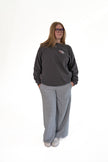 Woman wearing glasses and standing against a white background, wearing a charcoal gray crewneck sweatshirt with a small front chest graphic and wide-leg grey lounge pants, with her hands in her pockets