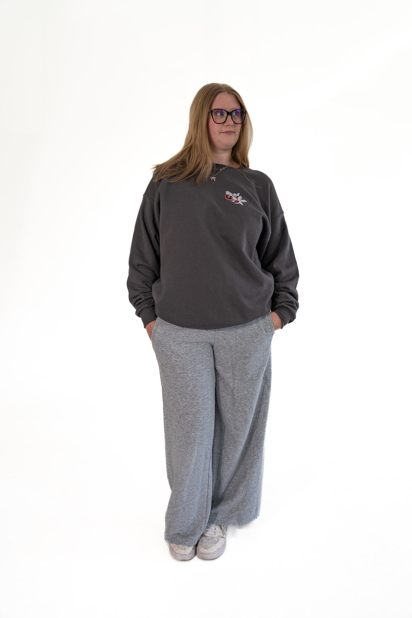 Woman wearing glasses and standing against a white background, wearing a charcoal gray crewneck sweatshirt with a small front chest graphic and wide-leg grey lounge pants, with her hands in her pockets