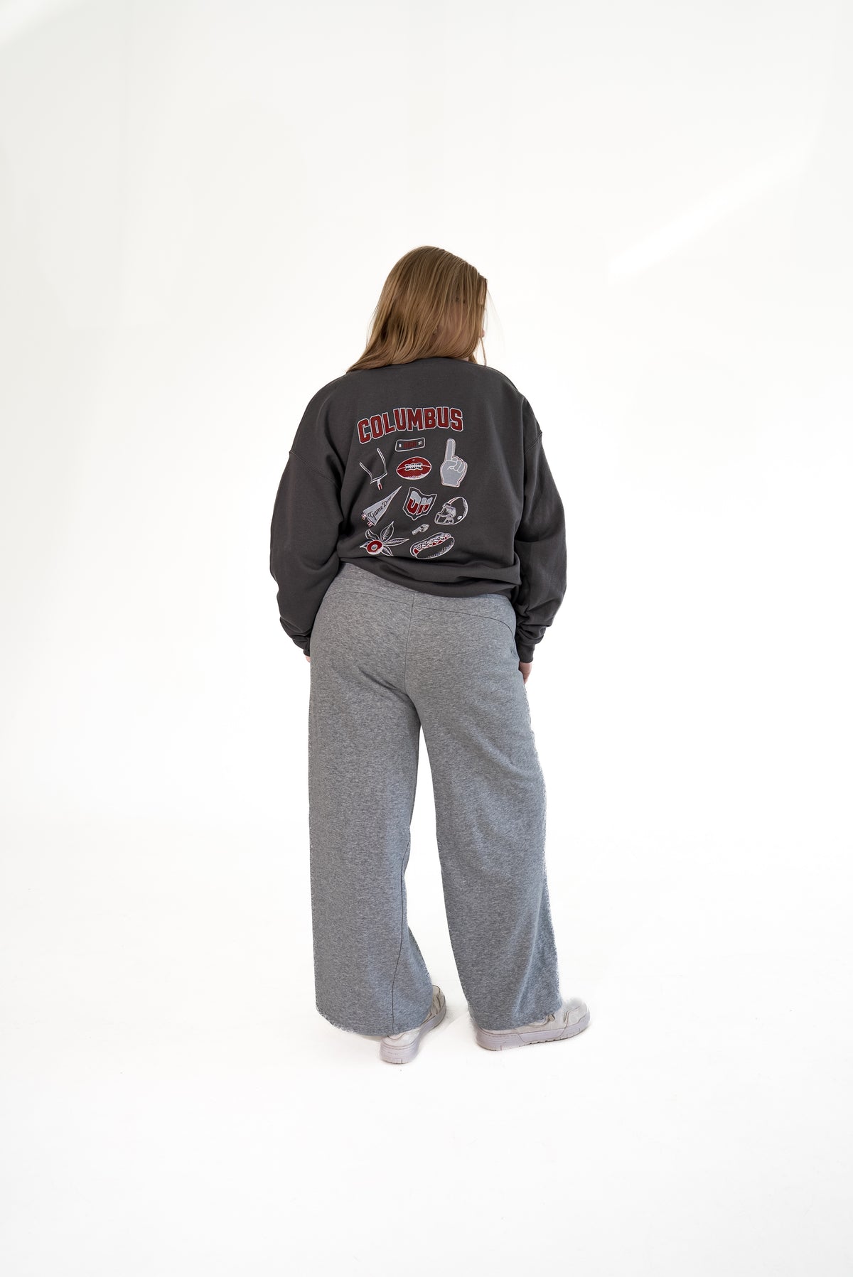 Woman standing against a white background from behind, wearing a charcoal gray crewneck sweatshirt with back game-day graphics, paired with wide-leg grey lounge pants.