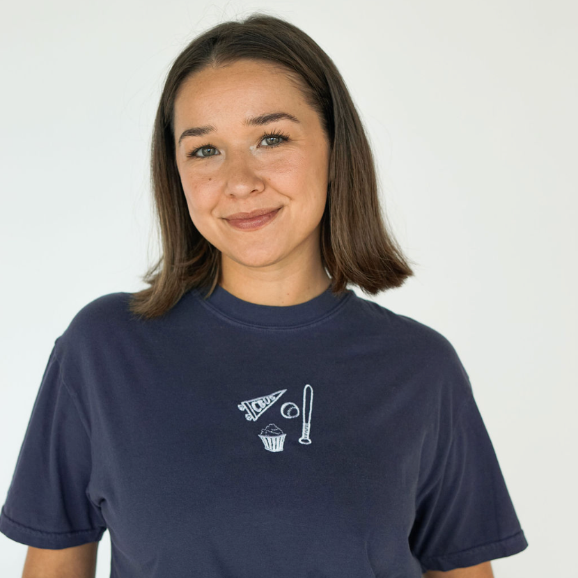 Model wearing a navy Columbus baseball tee embroidered with a CBUS pennant, baseball bat, baseball, and popcorn.