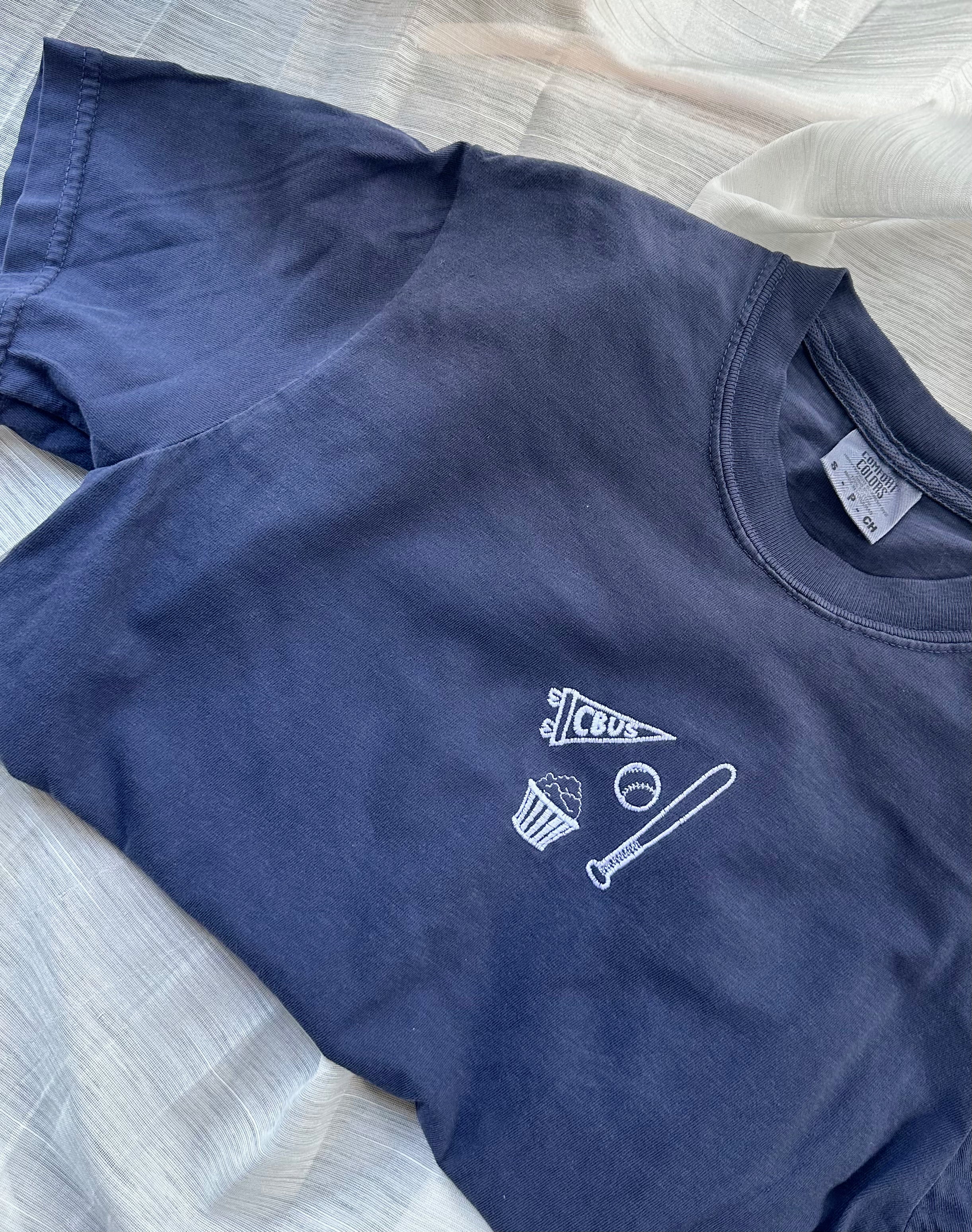 Flat lay of a navy Comfort Colors Columbus baseball T-shirt with embroidered CBUS pennant, baseball bat, baseball, and popcorn design.