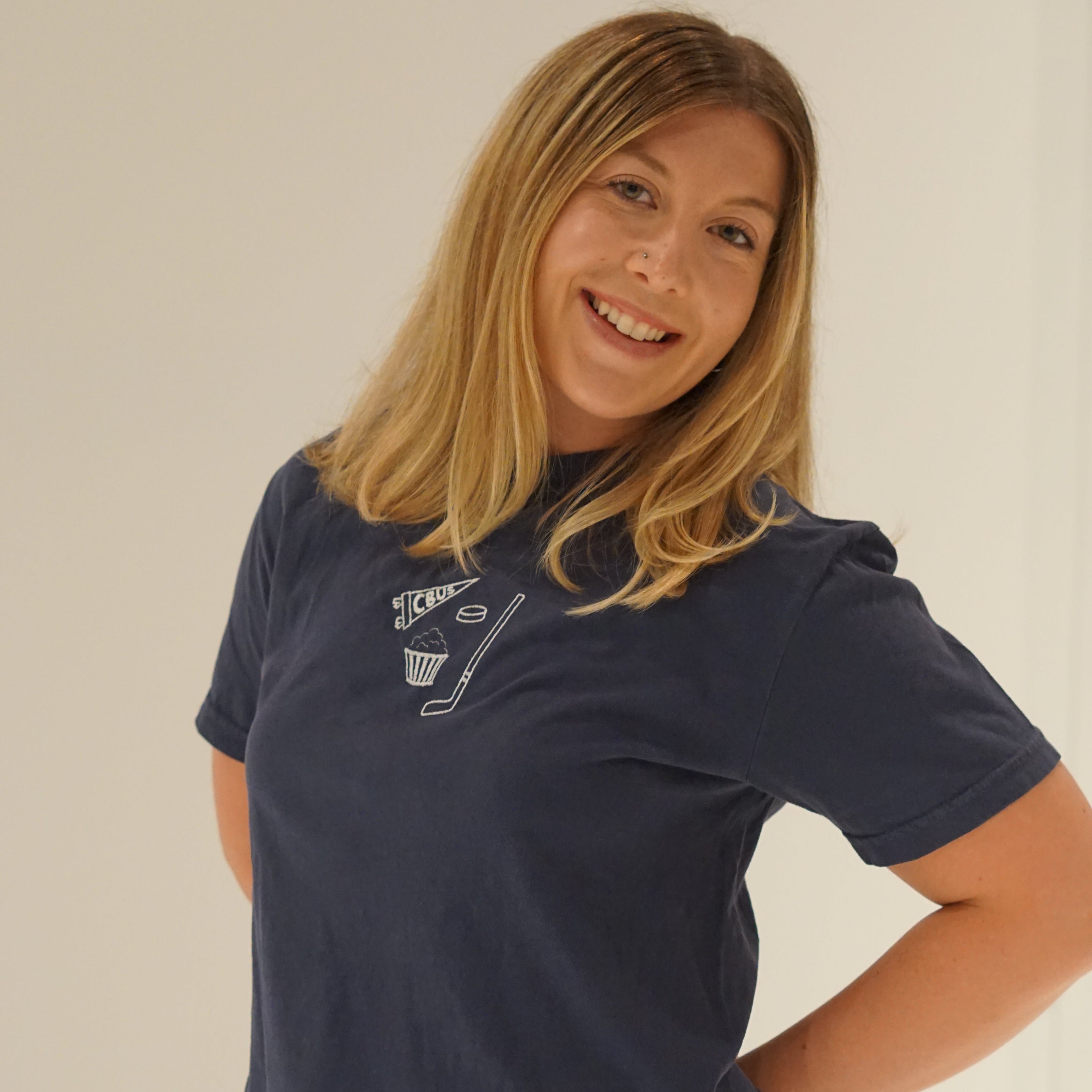 Woman wearing a navy, Comfort Colors Columbus hockey embroidered T-shirt featuring a CBUS pennant, hockey stick, puck, and popcorn design.
