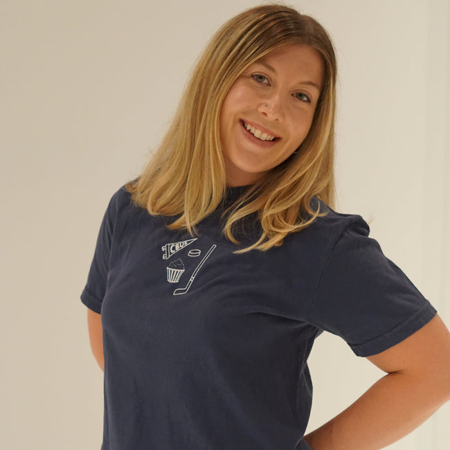 Woman wearing a navy, Comfort Colors Columbus hockey embroidered T-shirt featuring a CBUS pennant, hockey stick, puck, and popcorn design.
