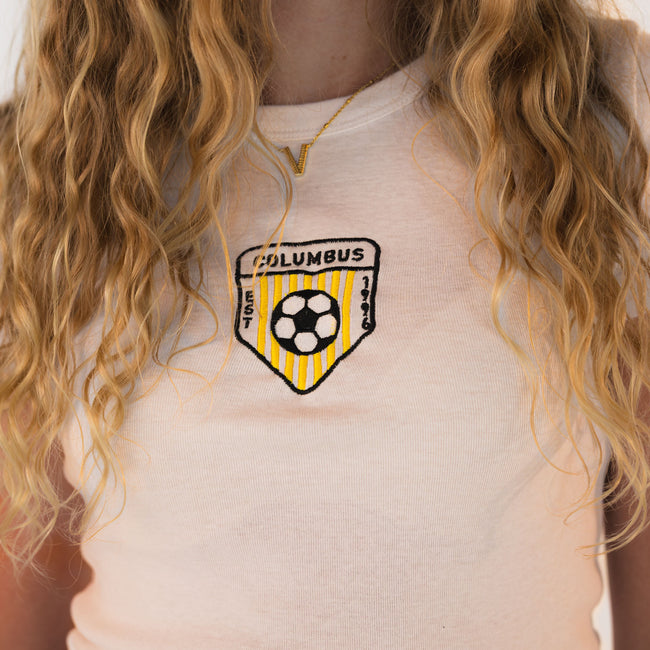 Close-up of the embroidered Columbus soccer crest on a peach women's baby tee.