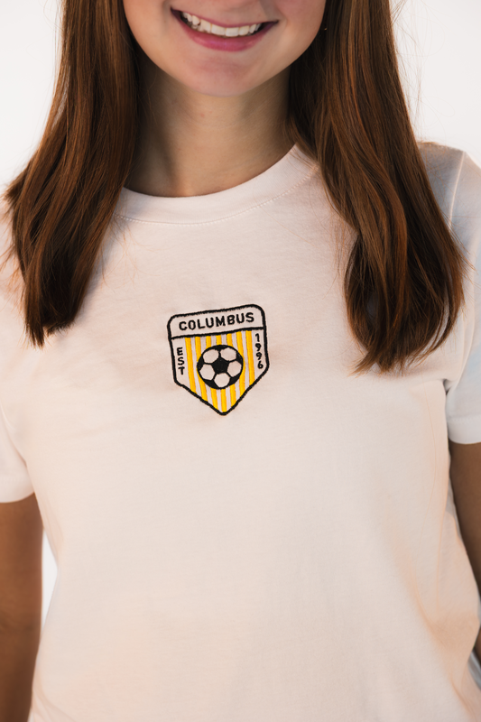Detailed embroidery of the Columbus soccer crest featuring black, white, and gold stitching on a white baby tee.