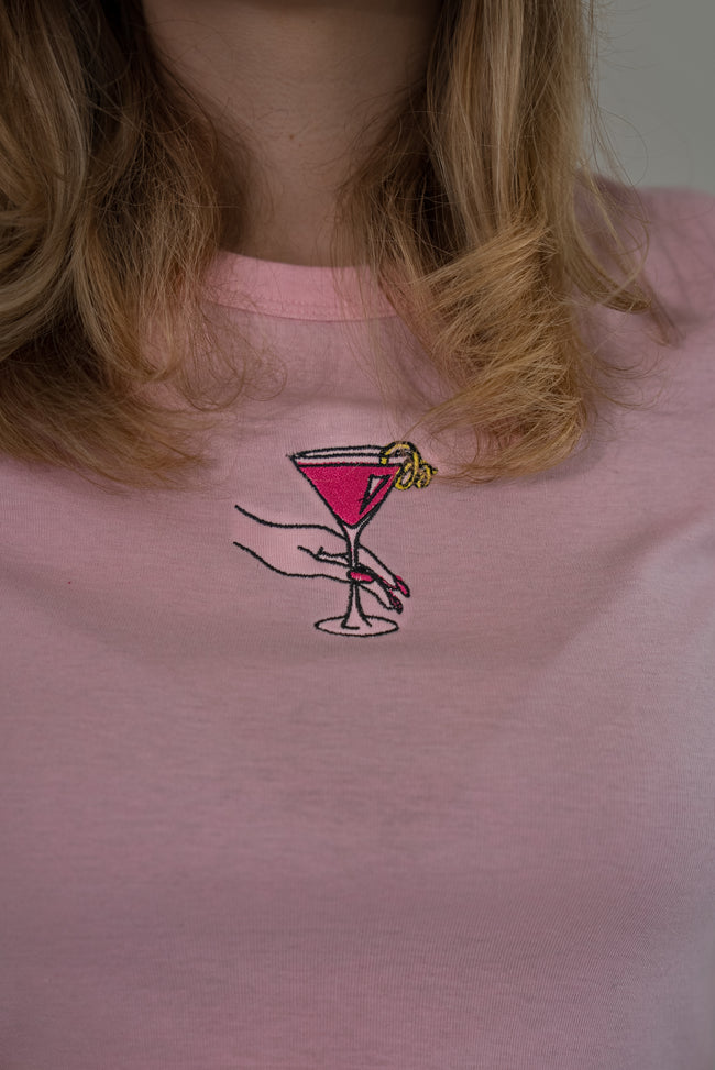 A close-up of a person wearing a light pink crewneck t-shirt with a small embroidered graphic of a hand holding a pink martini glass on the chest.