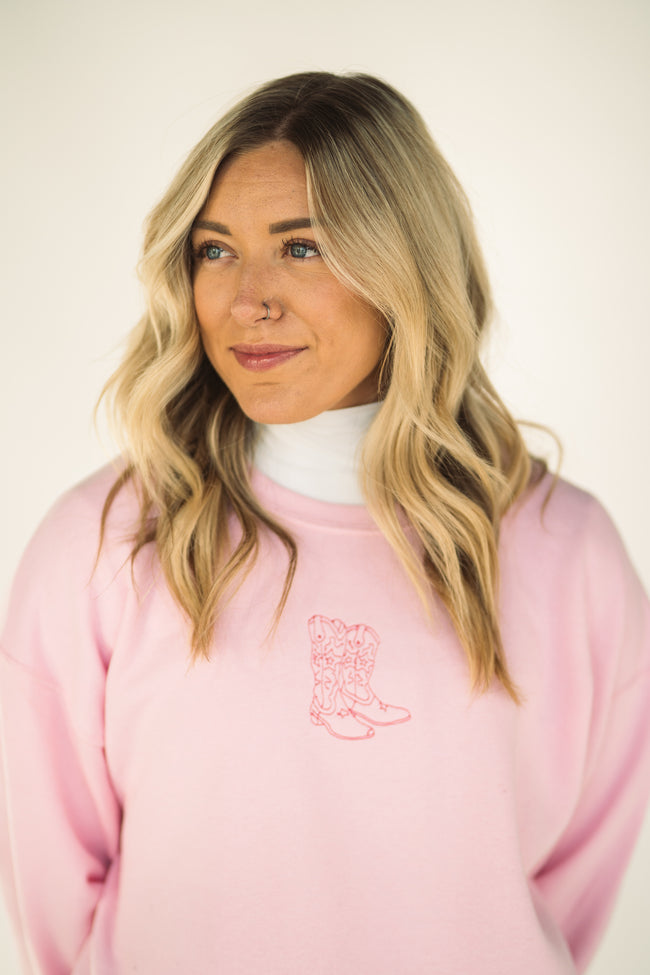 Woman wearing a light pink crewneck sweatshirt with an embroidered cowboy boot graphic on the chest, layered over a white mock neck top.