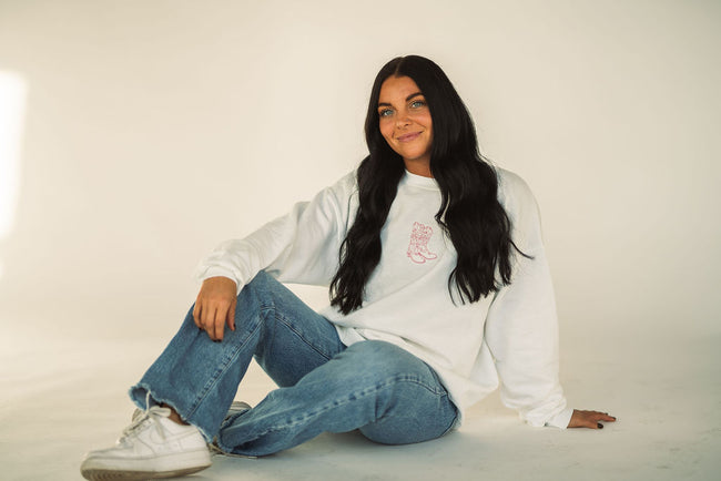 Woman sitting on the floor wearing a white crewneck sweatshirt with a pink embroidered cowboy boot graphic on the chest, styled with light wash jeans and white sneakers.