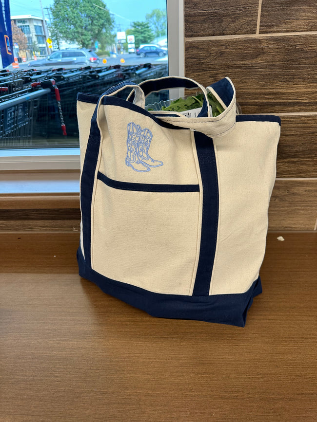 Cream canvas tote bag with navy trim embroidered with western cowgirl boots, displayed on a table.
