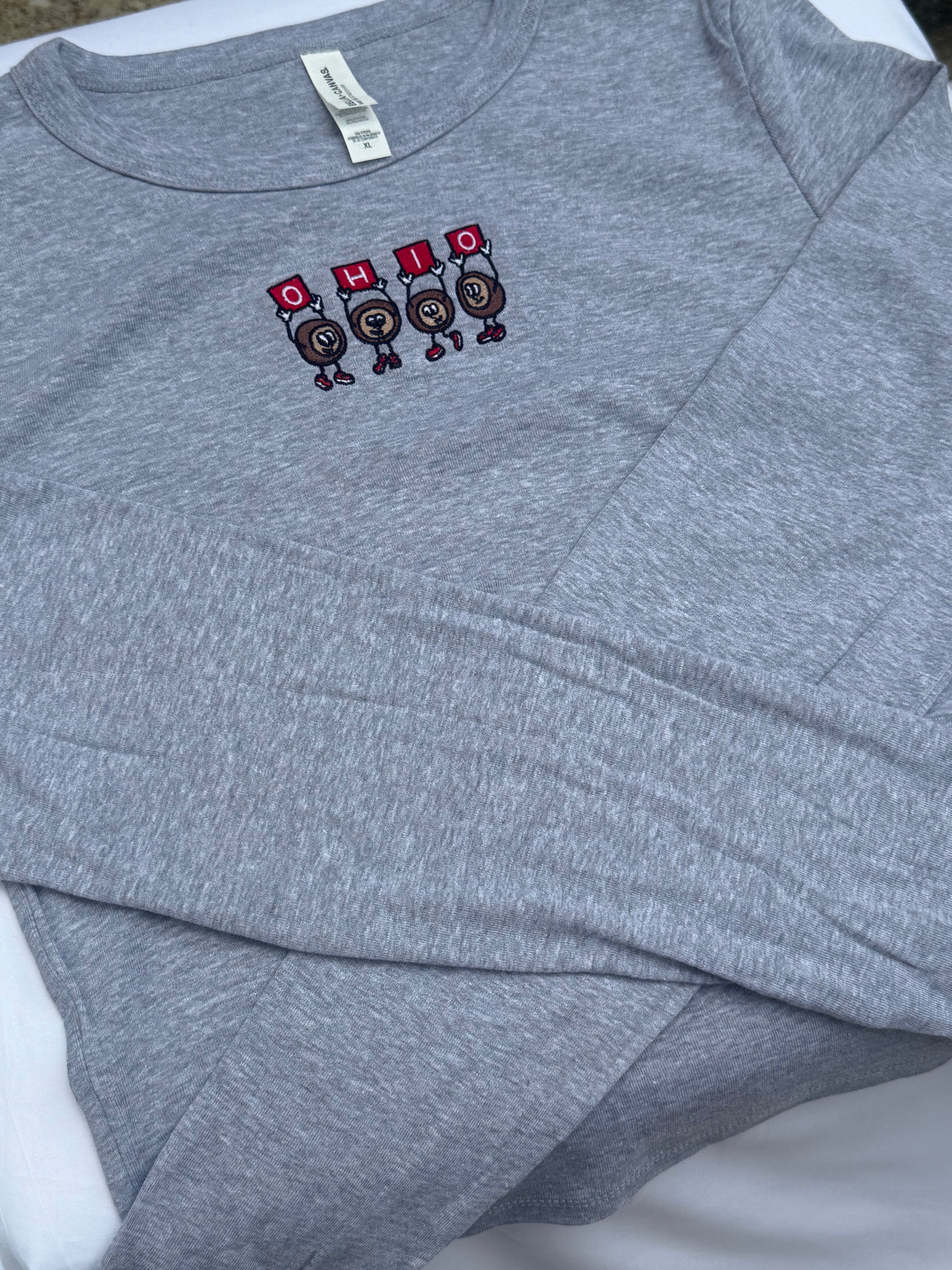 A folded heather gray long-sleeve shirt with a small chest graphic featuring four cartoon buckeyes holding red signs that spell 'OHIO'.